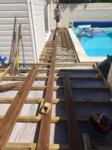 Un homme construit une terrasse en bois près d'une piscine, posant des lames sur une ossature en bois traitée reposant sur une toile géotextile.