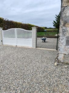Portail blanc à barreaux incurvés, devant une allée de gravier, à côté d'un pilier en pierre et d'une clôture grillagée où est posée une brouette.