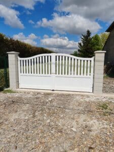 Portail battant blanc à claire-voie cintrée, encadré par deux piliers en blocs de pierre claire, sous un ciel bleu et nuageux.