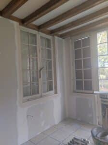 Intérieur en rénovation avec murs en plaques de plâtre jointoyées, fenêtres anciennes à petits carreaux, plafond à poutres apparentes et sol carrelé.