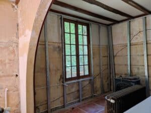 Rénovation intérieure : montage d'une ossature métallique (montants et rails) devant un mur ancien, une fenêtre à petits bois et un radiateur en fonte.