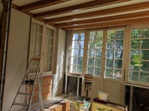 Chantier de rénovation : installation de plaques de plâtre et isolation Rockwool près d'une grande porte-fenêtre ancienne à petits carreaux donnant sur la verdure.