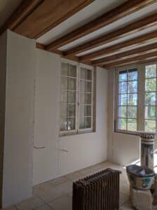 Intérieur en cours de rénovation avec plaques de plâtre fixées, poutres en bois apparentes, et fenêtres anciennes donnant sur des arbres verts.