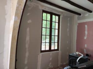 Intérieur en rénovation avec murs en plaques de plâtre jointoyées, une fenêtre en bois à petits carreaux et un poêle à bois noir.