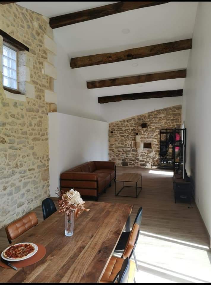 Maison rénovée : salon rustique chic poutres bois Salon moderne et rustique avec murs en pierre apparente, poutres en bois foncées, canapé en cuir marron, et grande table à manger en bois.