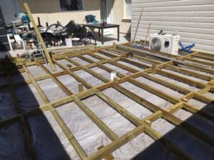 Construction d'une terrasse en bois : charpente de lambourdes vertes traitées posée sur une toile géotextile, avec outils Makita et climatiseur à proximité.