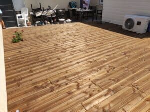 Grande terrasse en bois rainuré, ensoleillée, avec salon de jardin, climatiseur et décorations de nains de jardin.