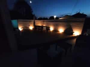 Terrasse en bois éclairée par des spots muraux chauds la nuit, mobilier de jardin sombre, ciel bleu nuit avec lune visible.