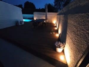 Terrasse en bois sombre éclairée par des spots muraux chauds la nuit, près d'une piscine bleue et d'un climatiseur extérieur.
