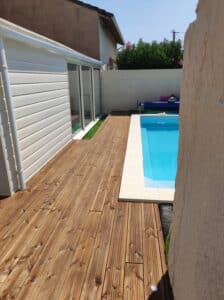 Terrasse en bois clair longeant une piscine rectangulaire et une annexe avec revêtement vinyle blanc sous un ciel bleu.