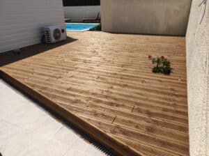 Grande terrasse en bois clair rainuré près d'une piscine, avec une unité extérieure de climatisation et une petite plante à fleurs rouges.