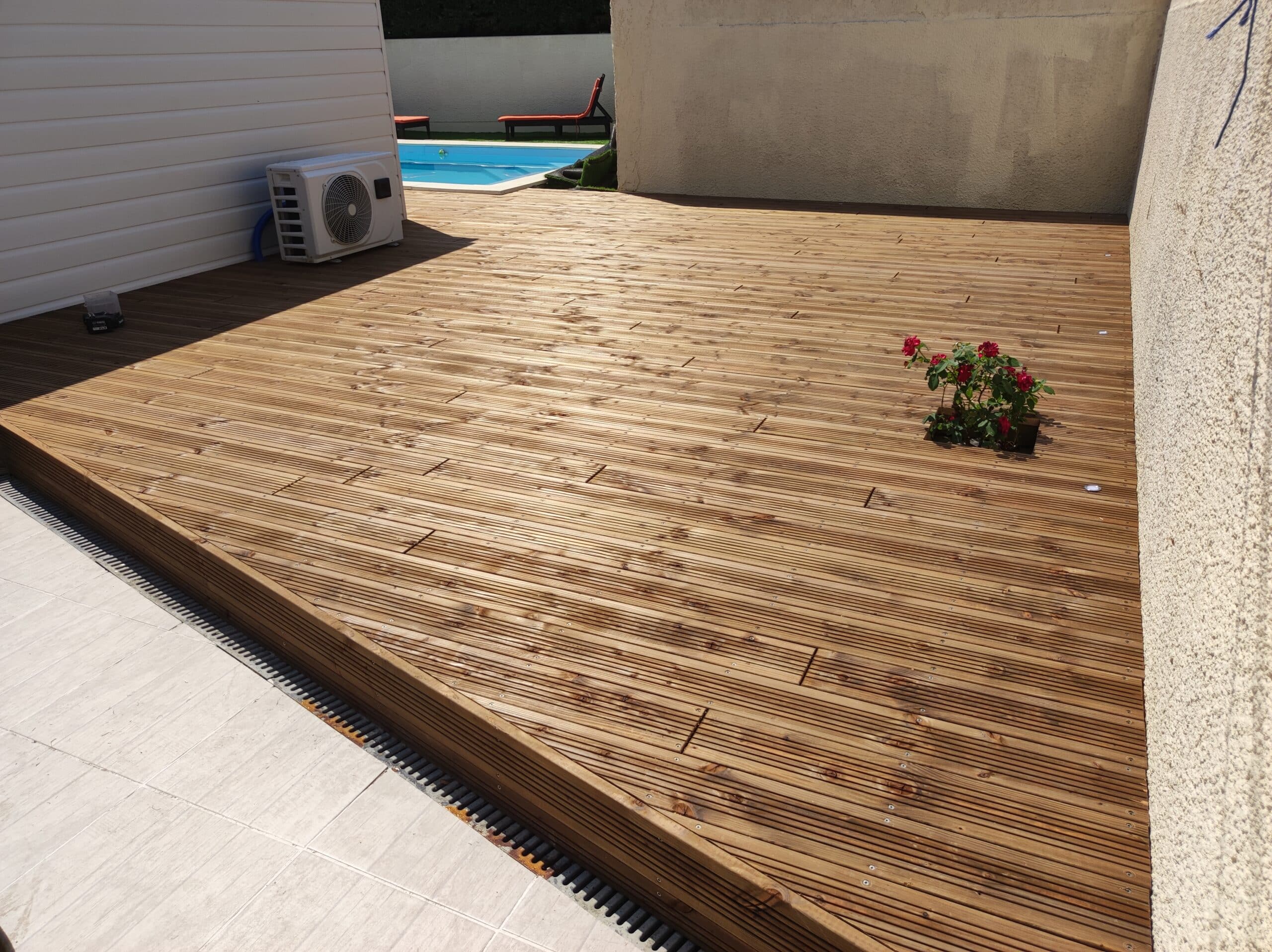 Grande terrasse en bois clair rainuré près d'une piscine, avec une unité extérieure de climatisation et une petite plante à fleurs rouges.